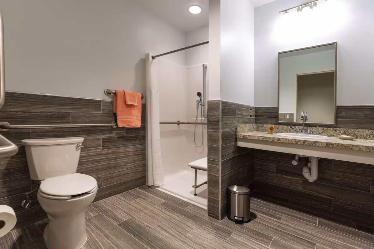A modern bathroom with wood-patterned tile flooring and half-wall tiles. It features a white toilet with grab bars, a walk-in shower with a white curtain and a built-in seat, a granite countertop with a sink, a large mirror, and a small trash can underneath the counter. An orange towel hangs on a grab bar next to the toilet.