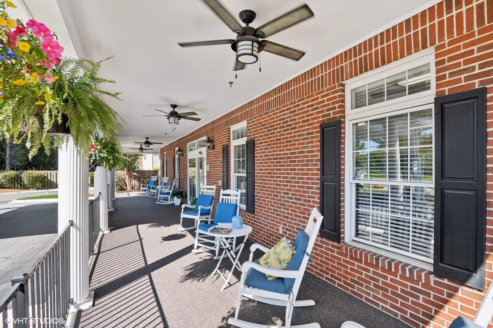 Covered brick-front porch with rocking chairs, ceiling fans, and hanging plants at a senior living facility entrance.