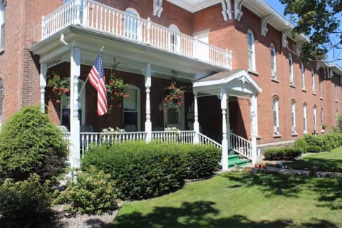 Brick senior living facility with a white porch, railing, American flag, and manicured lawn.