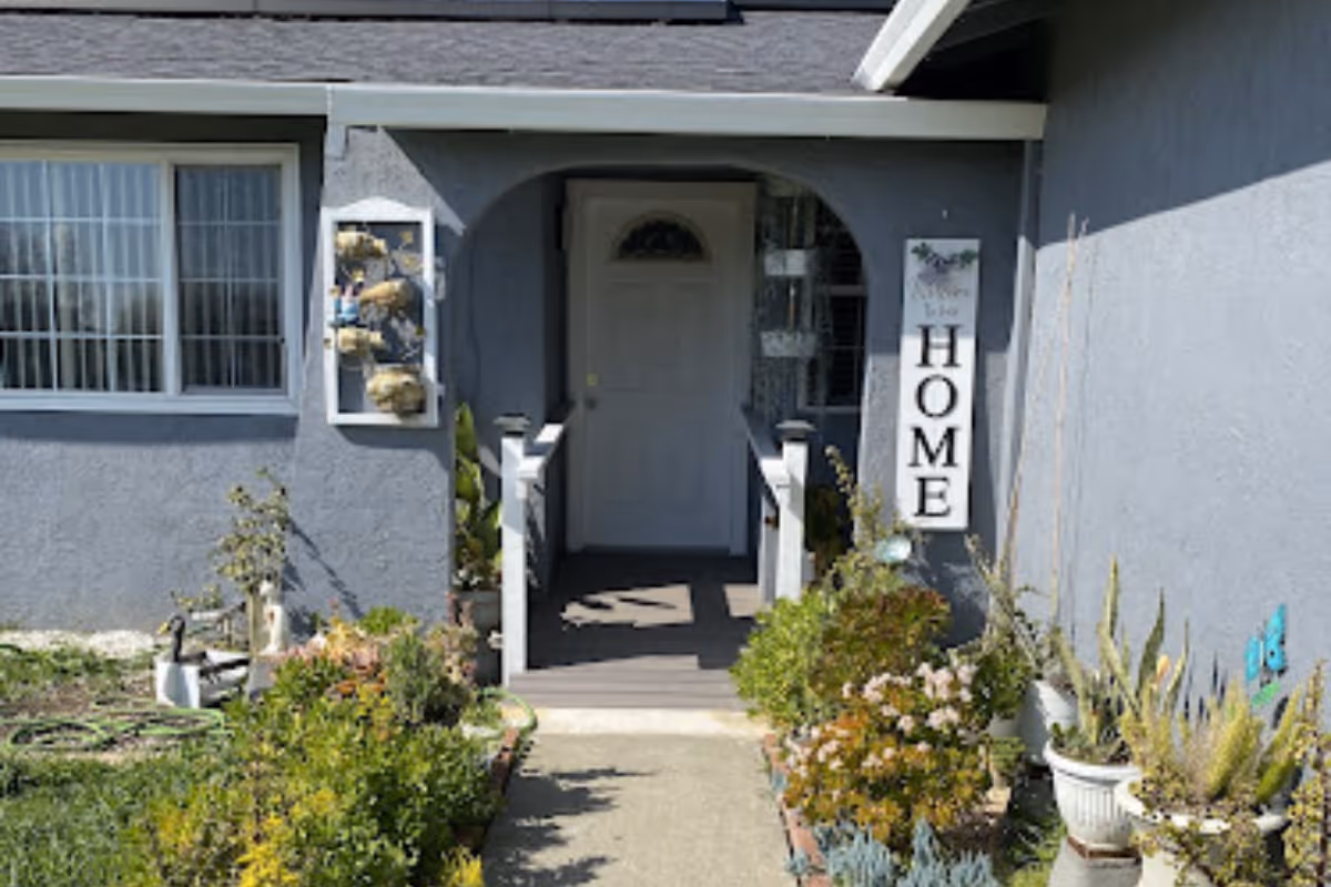 Front entrance of a residential building with a small porch, a white door, and a sign that reads 'Welcome to our HOME'. There are various plants and flowers in pots and garden beds along the walkway leading to the door.