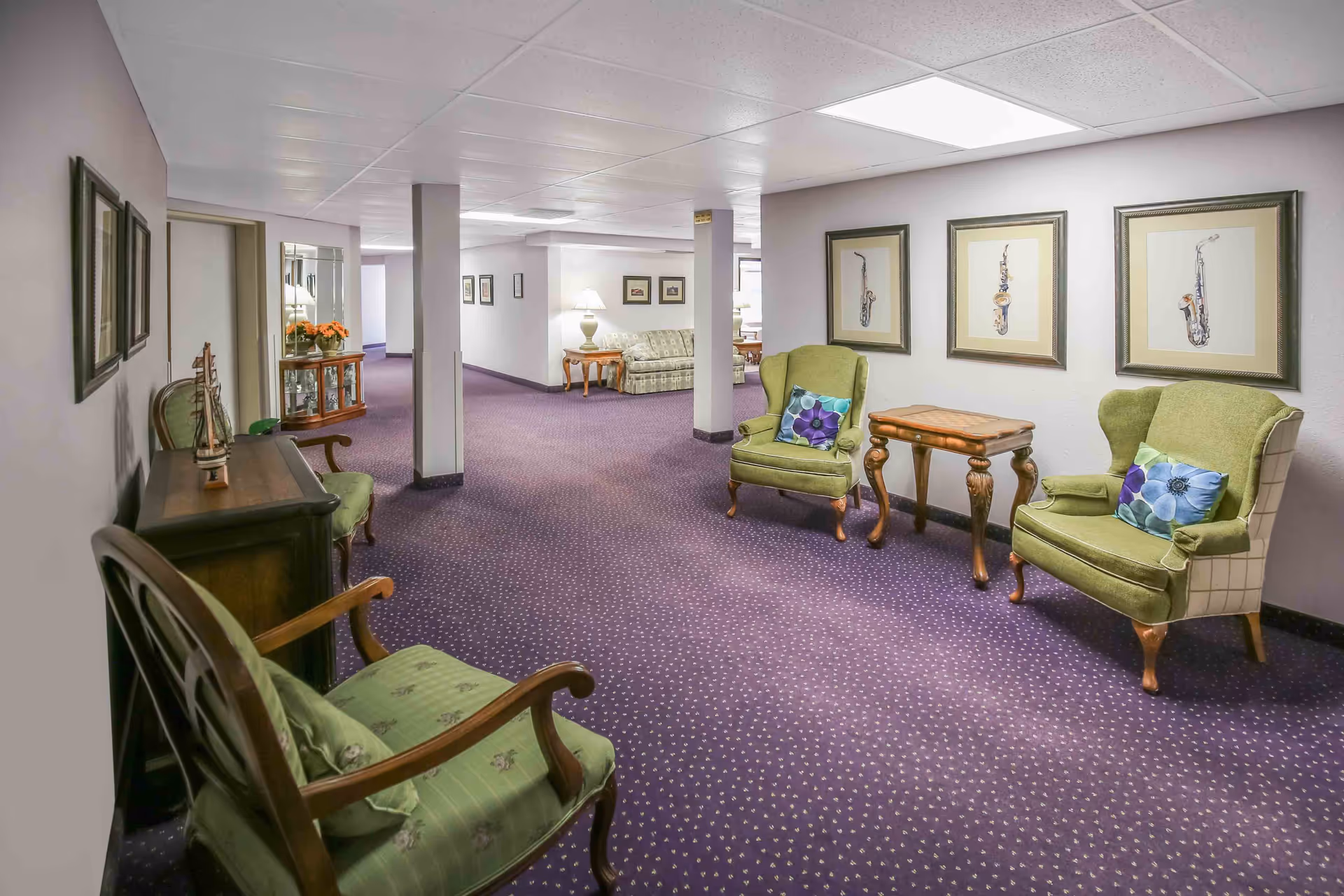 Carpeted communal seating area with green armchairs, small tables, framed artwork, and a purple patterned carpet.