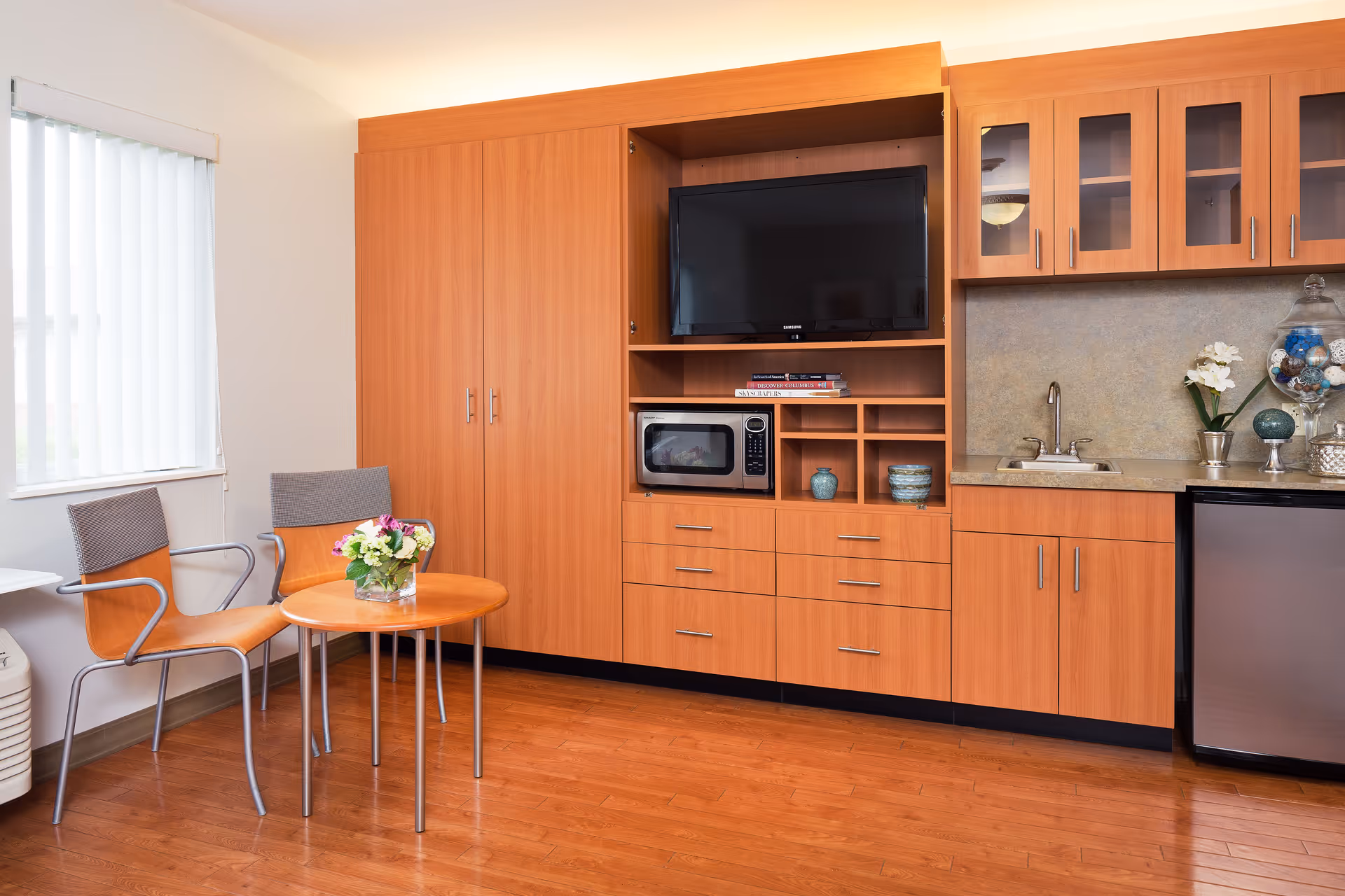 A cozy interior room featuring wooden cabinetry with a mounted flat-screen TV, a microwave, and a small sink area. There are two modern chairs with metal frames and wooden seats around a small round wooden table with a floral arrangement. A window with vertical blinds allows natural light into the room.