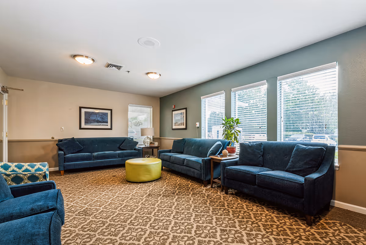 Communal living room with teal sofas, patterned carpet, a round green ottoman, side tables and windows along one wall.