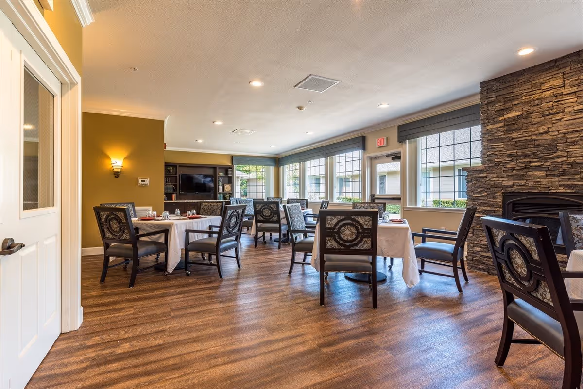 A bright dining room with several tables covered with white tablecloths and set with glasses and napkins. The room has wooden flooring, large windows with gray blinds, a stone fireplace on the right, and a TV mounted on the far wall. The walls are painted in warm tones, and there are decorative chairs around the tables.