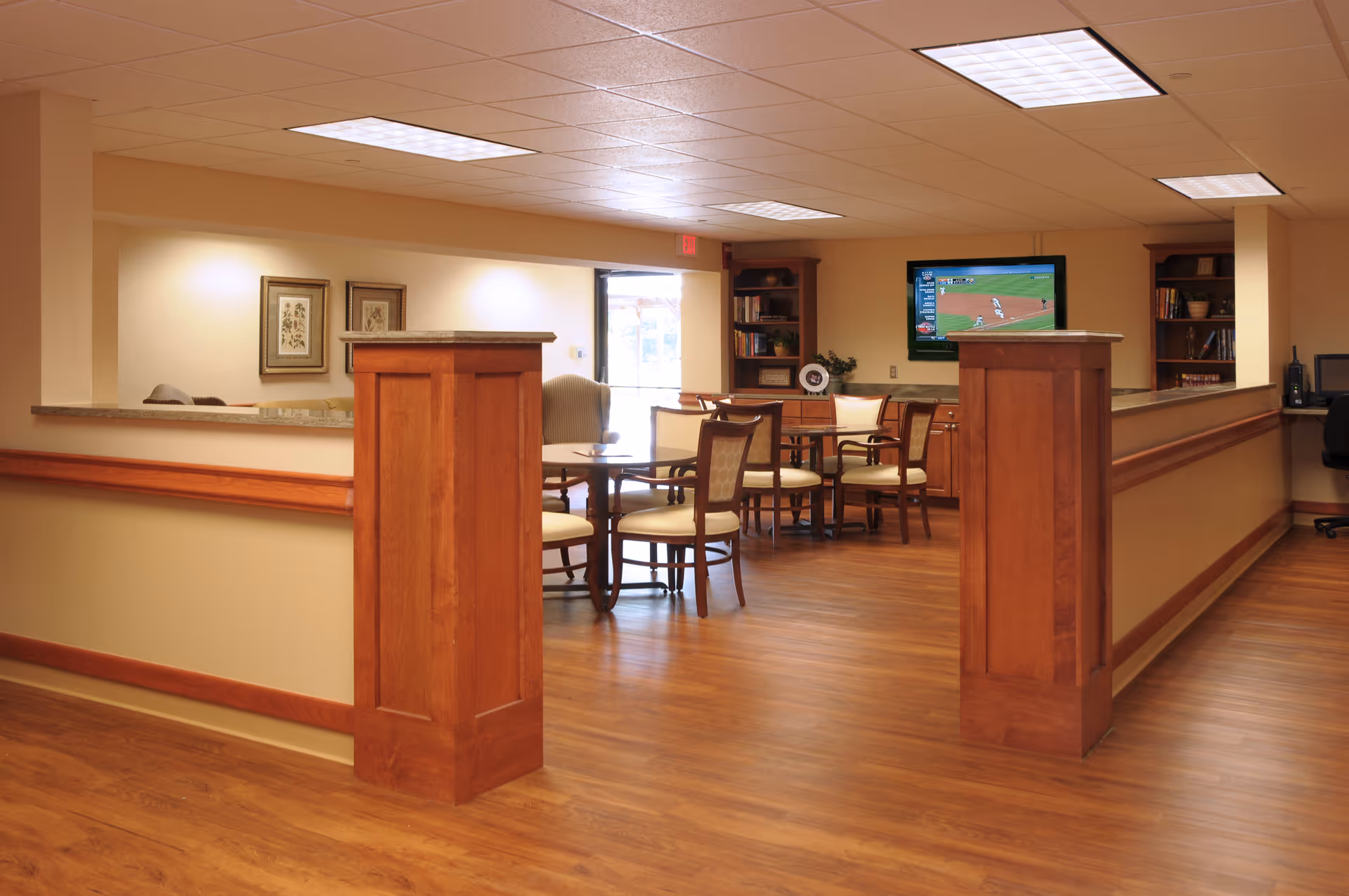 Bright communal interior space with wood paneling, round tables and chairs, bookshelves, and a wall-mounted TV.