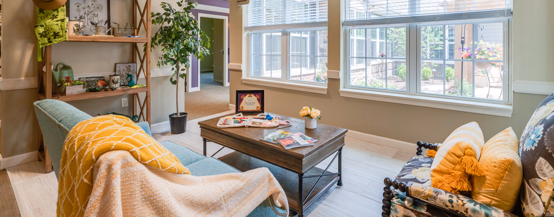 A cozy living room area with a light blue sofa draped with a yellow and white blanket, a wooden coffee table with magazines and a small vase of flowers, and a floral-patterned armchair with yellow pillows. There is a tall potted plant next to a wooden shelving unit holding decorative items. Large windows let in natural light and show a view of an outdoor garden area.