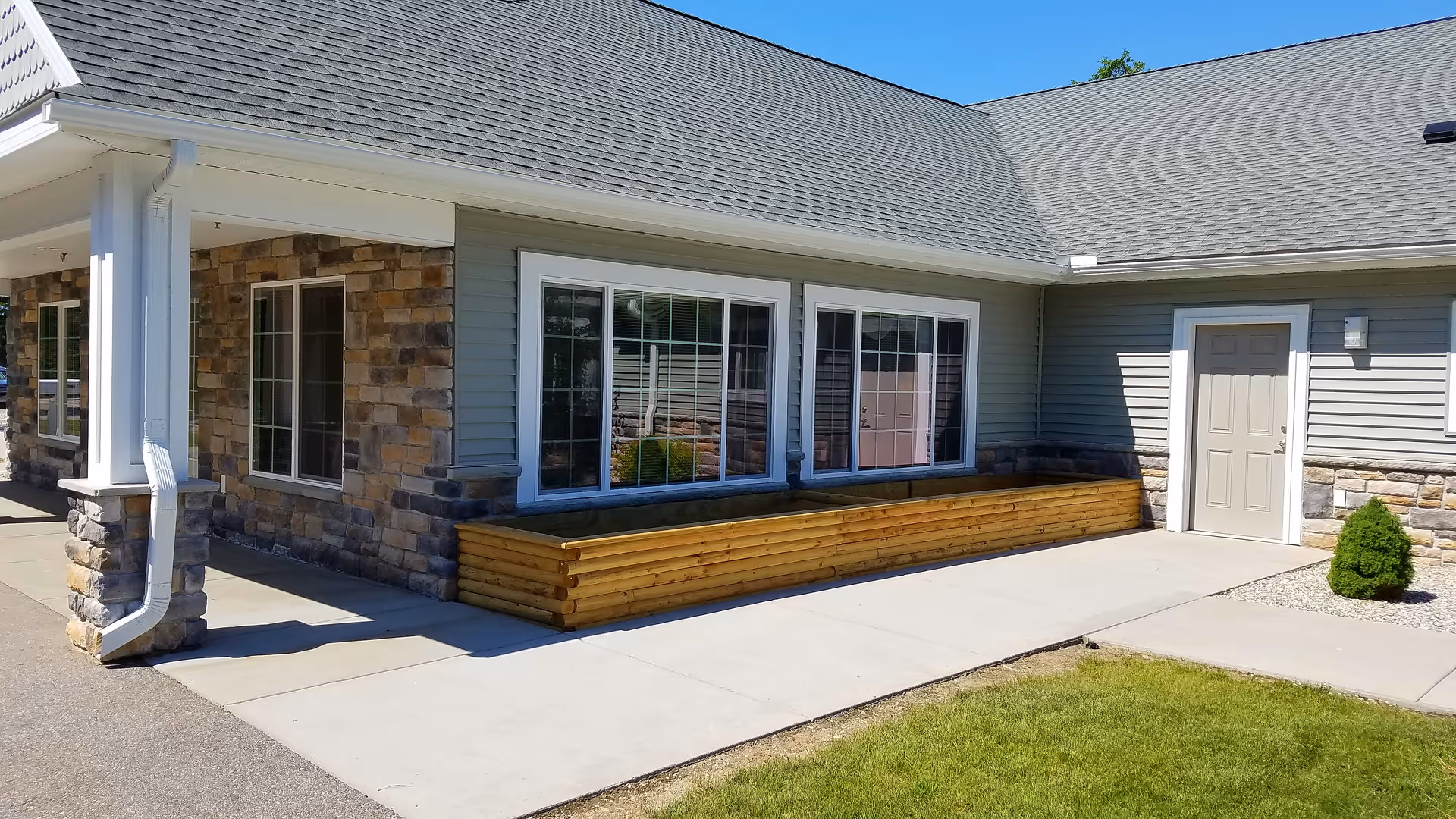 Exterior front entrance of a single-story assisted living building with stone and siding facade, large windows, a wooden planter box, and a concrete walkway.
