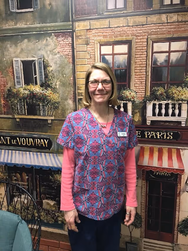 A woman wearing glasses and a colorful patterned scrub top over a pink long-sleeve shirt stands smiling in front of a mural depicting a European-style building facade with windows, flower boxes, and awnings.