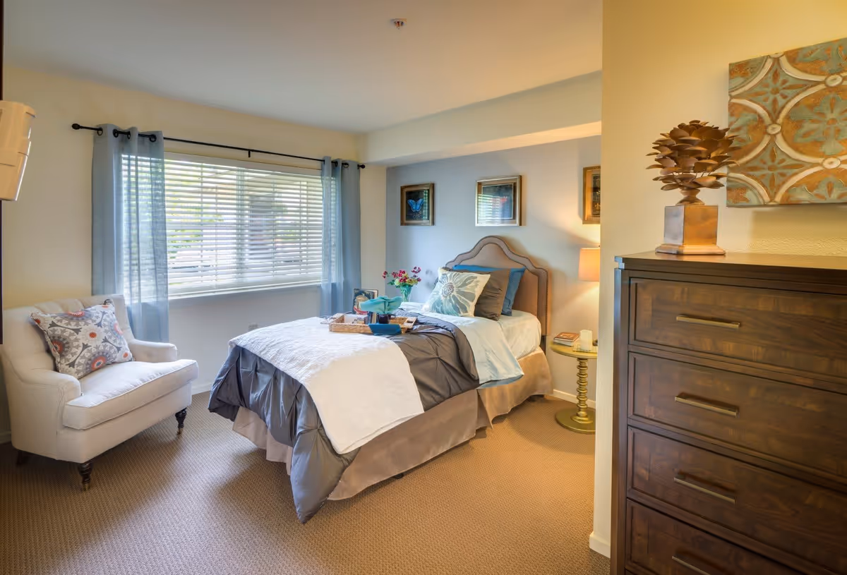 A cozy bedroom with a single bed dressed in gray and white bedding, a cushioned armchair with a patterned pillow, a wooden dresser with decorative items on top, and a window with sheer blue curtains letting in natural light.