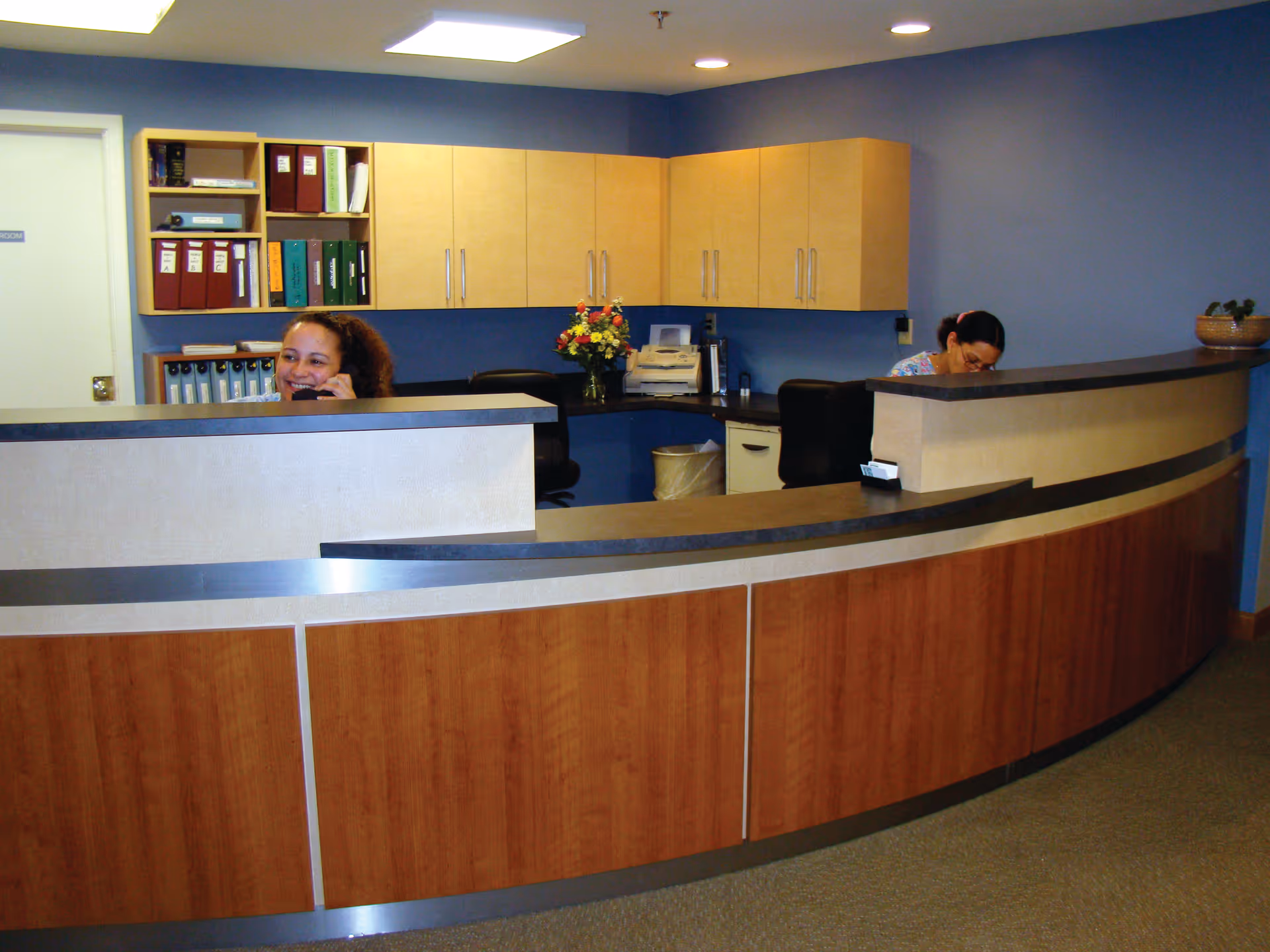 Reception area with a curved wooden desk and two women working behind it. One woman is smiling and talking on the phone, while the other is focused on paperwork. The background features blue walls, wooden cabinets, shelves with binders, and a bouquet of flowers on the counter.