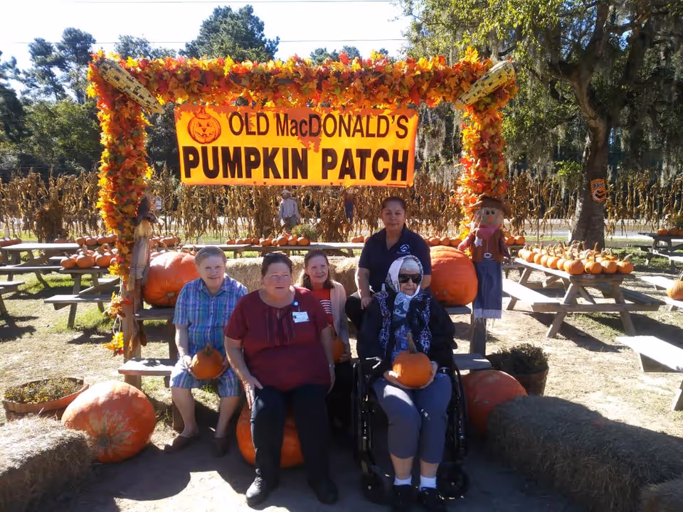 A group of five people, including elderly individuals and a caregiver, posing at a pumpkin patch decorated with autumn leaves and a sign that reads 'OLD MacDONALD'S PUMPKIN PATCH'. They are seated on hay bales and holding pumpkins, with picnic tables and more pumpkins visible in the background under a sunny sky.