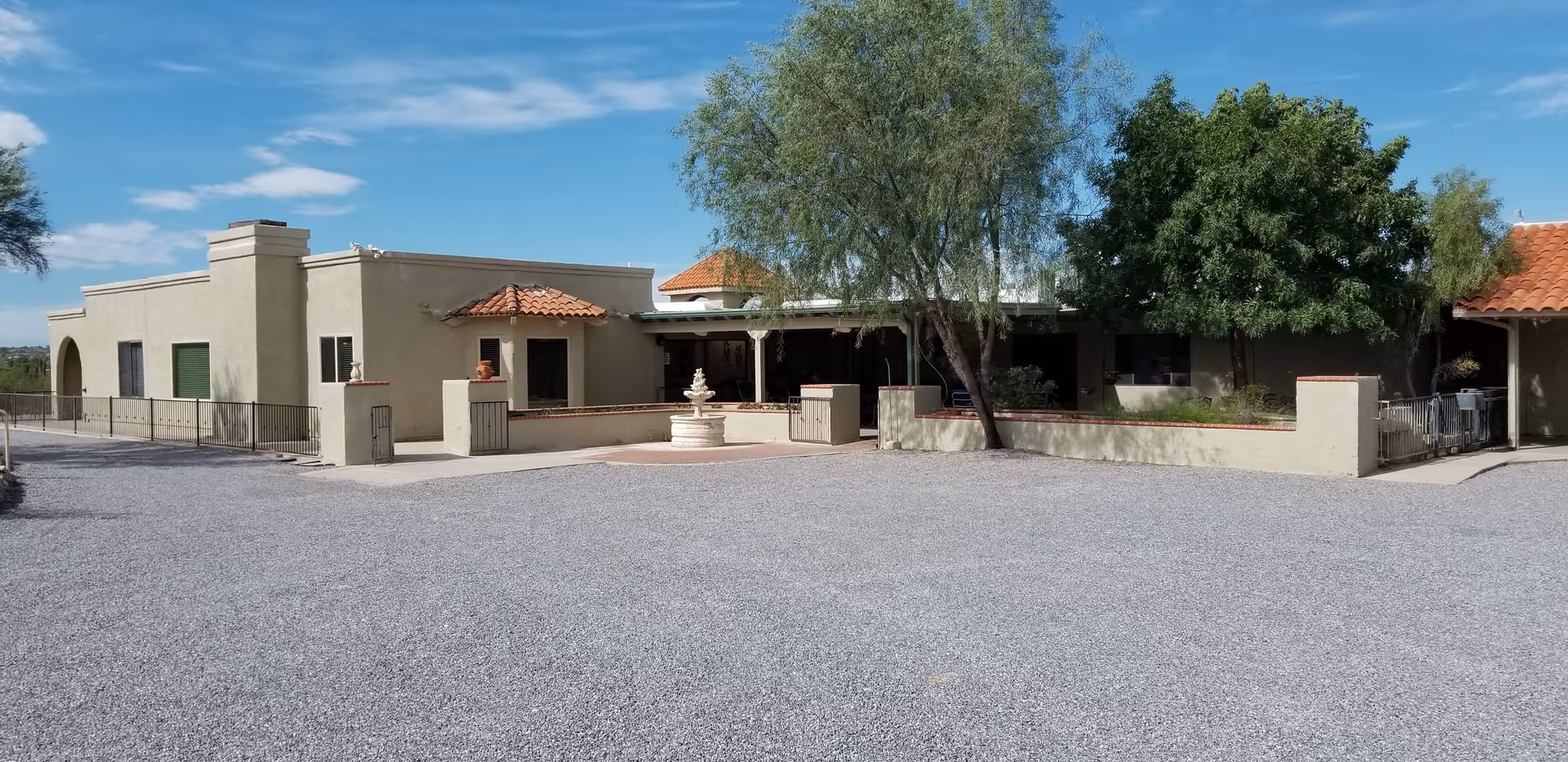 Exterior view of a single-story building with beige stucco walls and red tile roofing, surrounded by trees and a gravel driveway. The building has a small courtyard with a white fountain in front.