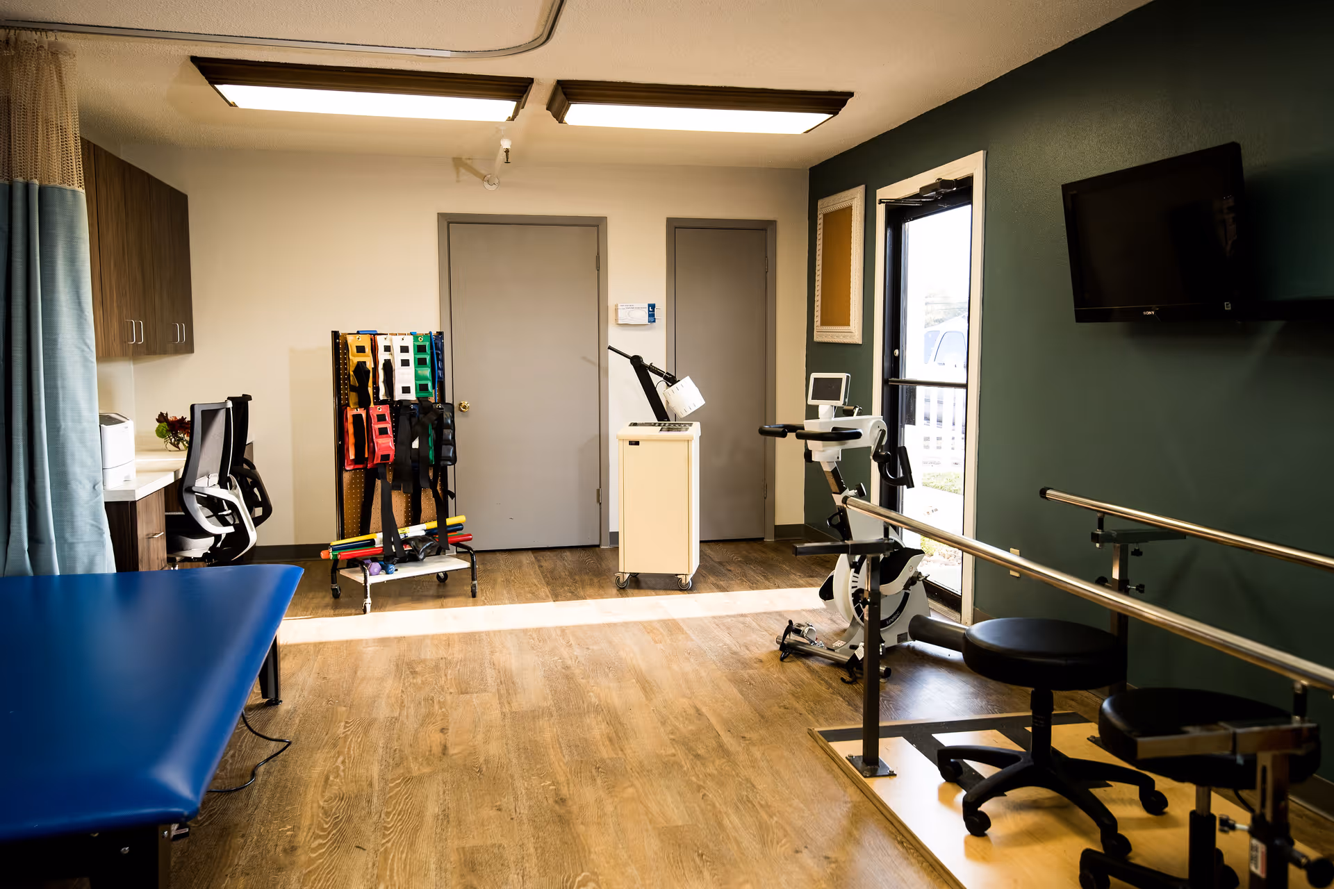 A rehabilitation therapy room with parallel bars, exercise equipment, a treatment table, and storage cabinets.
