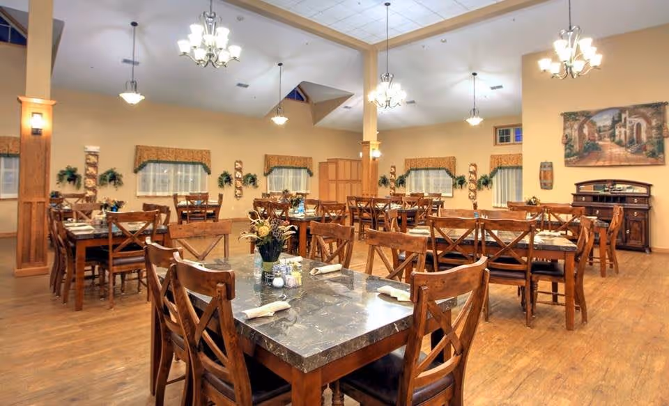 A spacious dining room with multiple wooden tables and chairs arranged neatly. Each table is set with napkins, salt and pepper shakers, and a small flower arrangement. The room has warm lighting from chandeliers hanging from the ceiling, wood flooring, and windows with valances. Decorative plants and wall art adorn the walls, creating a welcoming atmosphere.
