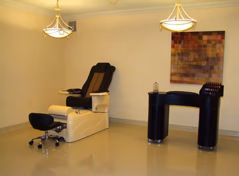 A clean and simple nail salon room featuring a beige pedicure chair with a black footrest, a black rolling stool, a black manicure table with nail polish bottles on top, and a colorful abstract painting on the wall. Two ceiling lights illuminate the room.