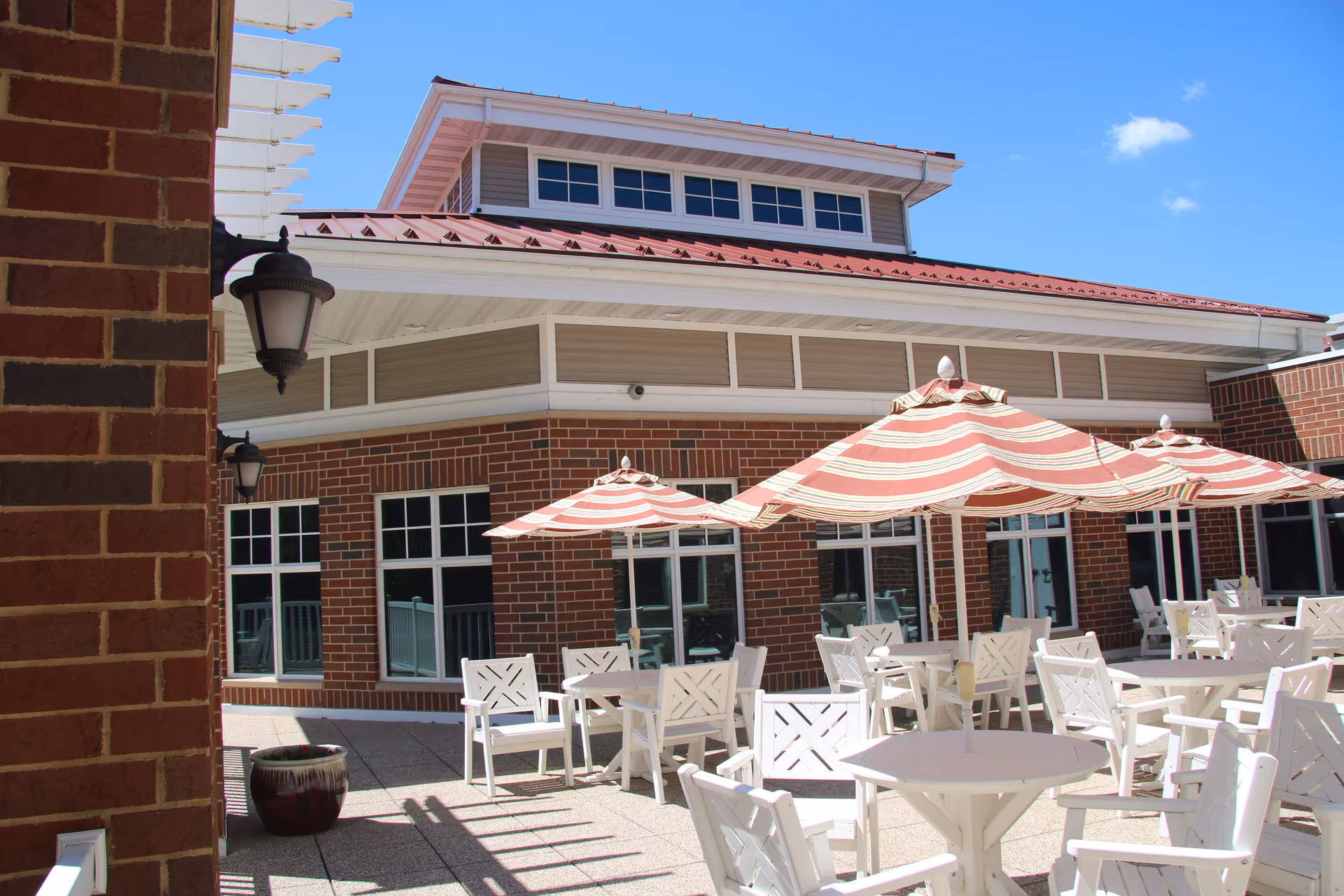 Outdoor patio area at Wittenberg Village: Lutheran Life Communities with white tables and chairs, and red and white striped umbrellas. The building has red brick walls and multiple windows under a red metal roof with a clear blue sky above.