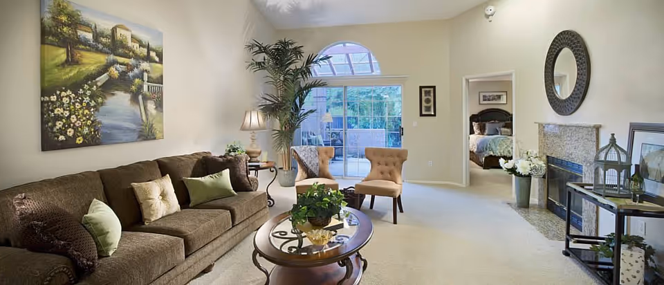 A cozy living room with a brown sofa adorned with green and beige pillows, a glass coffee table with a plant centerpiece, two beige upholstered chairs near a large arched window, and a fireplace with a round mirror above it. A bedroom with a bed is visible through an open doorway.