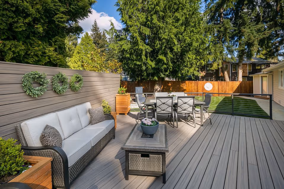 Outdoor patio area with a cushioned sofa, a coffee table with a flower pot, a dining table with six chairs, surrounded by wooden fencing and tall green trees under a blue sky with some clouds.