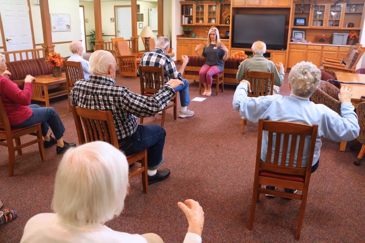 A group of elderly people seated on wooden chairs in a common room participating in a seated exercise session led by a woman in colorful leggings. The room has carpeted flooring, wooden furniture, and a large built-in wooden cabinet with a television.