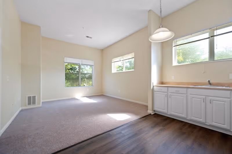 Empty room with beige walls and carpeted floor on the left side, and a kitchen area with white cabinets, a countertop, and a sink on the right side. Two windows allow natural light to enter the space, and a hanging light fixture is above the kitchen area.