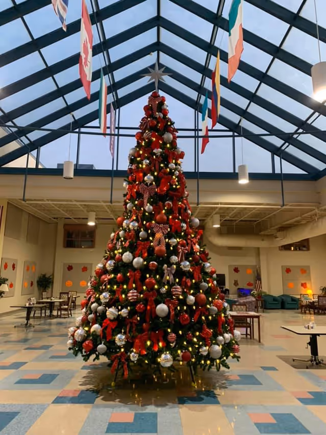 A large, decorated Christmas tree with red and silver ornaments and ribbons stands in the center of a spacious room with a high glass ceiling. Flags hang from the ceiling beams. The room has tables, chairs, and couches arranged around the perimeter, with festive decorations on the walls.