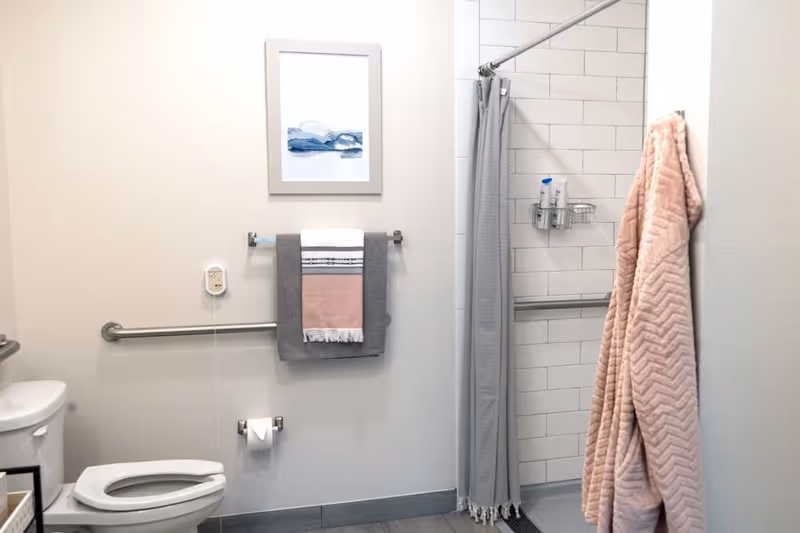 A clean and accessible bathroom with a toilet, grab bars, a towel rack holding folded towels, a shower area with a gray curtain, and a pink textured bathrobe hanging on the wall. There is also a framed abstract artwork on the wall above the towel rack.