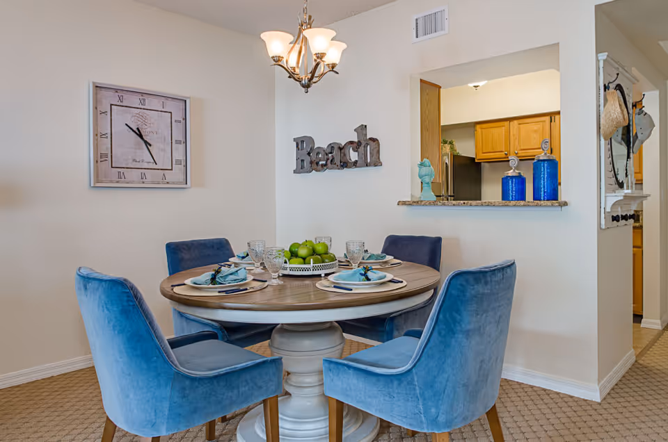 Round dining table set for four with blue upholstered chairs in a bright dining area featuring a wall clock, a 'Beach' sign, and a pass-through to the kitchen.