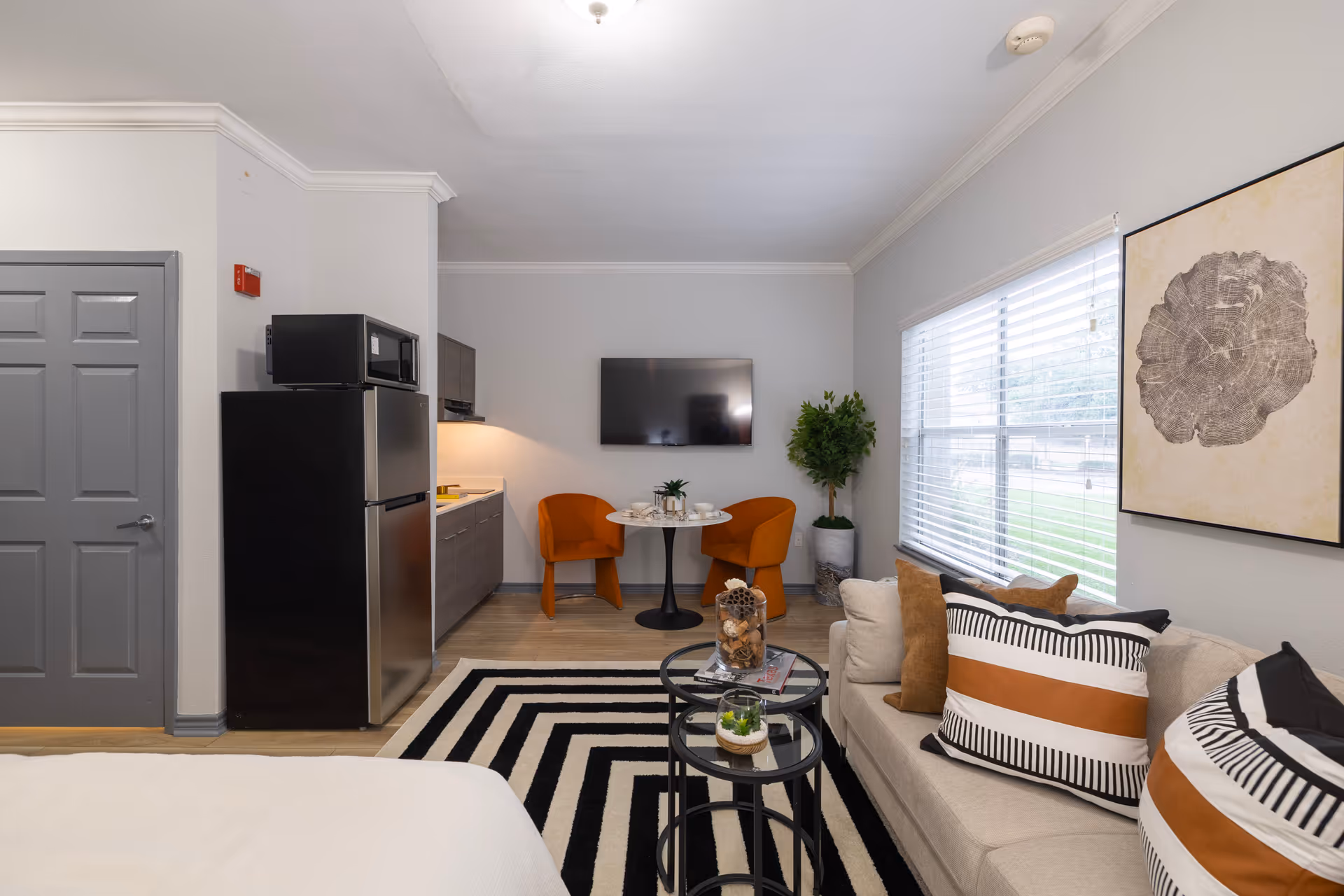 A modern studio apartment interior with a small kitchen area featuring a refrigerator and microwave, a round dining table with two orange chairs, a wall-mounted flat screen TV, a beige couch with decorative pillows, a large window with blinds, and a black and white striped rug on the floor.