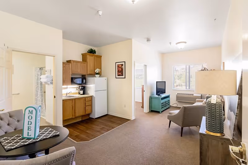 Interior view of a senior living apartment at The Chesterley featuring a small kitchen area with wooden cabinets, a white refrigerator, microwave, and coffee maker. Adjacent to the kitchen is a living area with a TV on a teal stand, a beige armchair, a sofa, and a lamp on a side table. A round dining table with two chairs is visible in the foreground, and a bathroom entrance is seen to the left with towels hanging on the door.