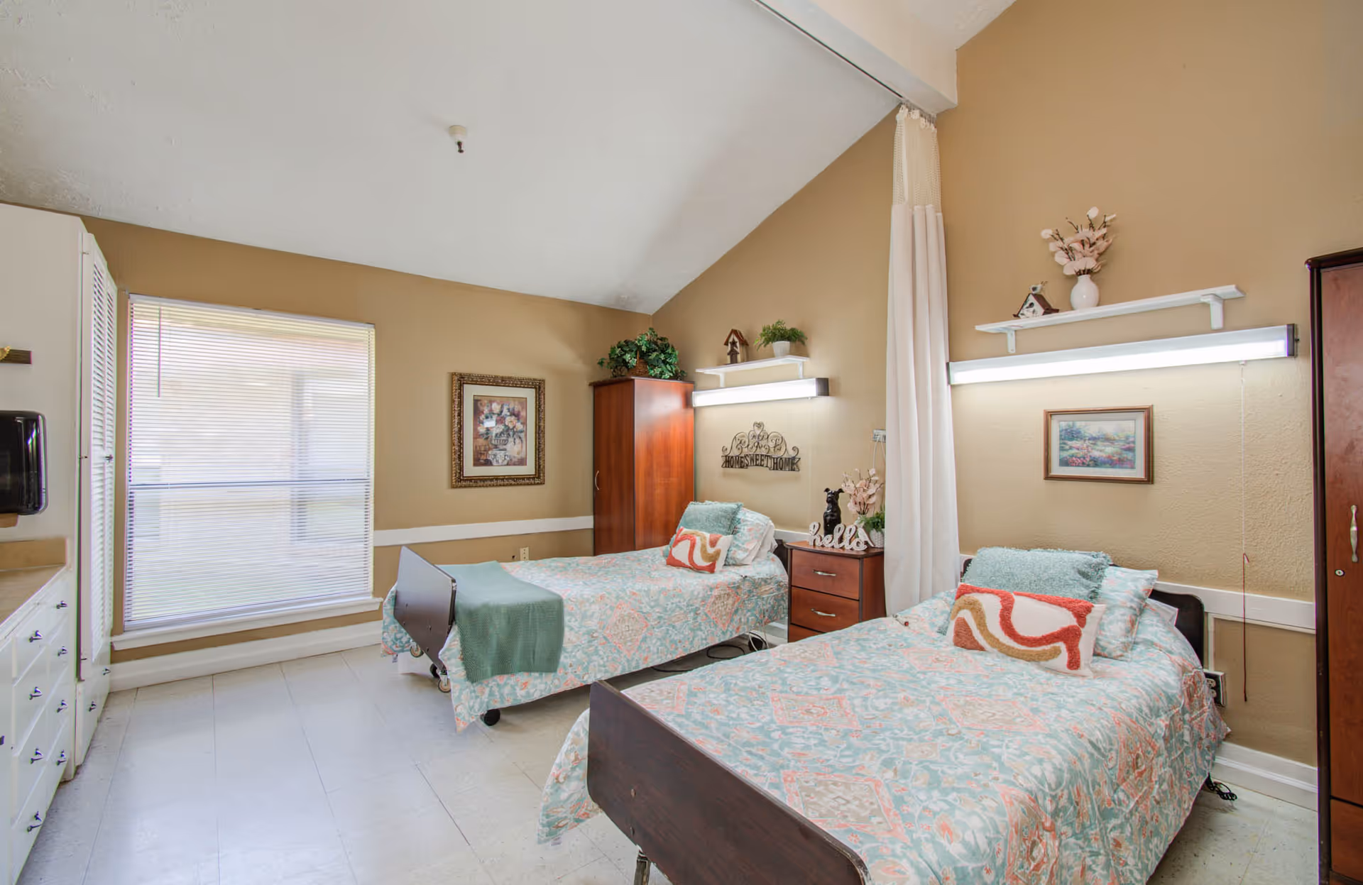 A nursing facility bedroom with two single beds, each with patterned bedspreads and decorative pillows. The room has beige walls, a large window with blinds, wooden wardrobes, a nightstand with decorative items, and wall-mounted lights above each bed.