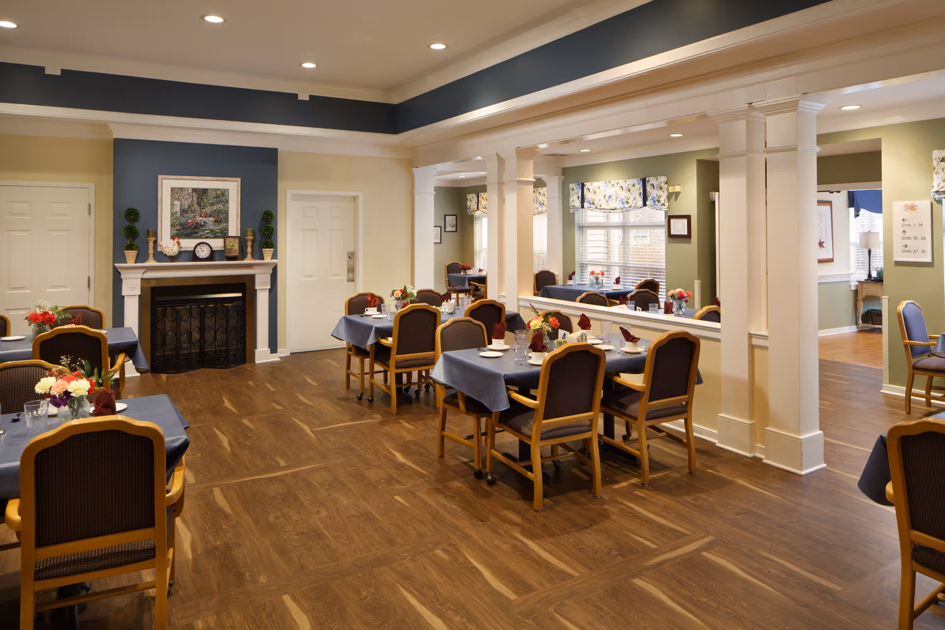 A dining room in a senior living facility with several tables covered in blue tablecloths, each set with plates, glasses, and napkins. The room features wooden flooring, a fireplace with decorative items on the mantel, and large windows with floral valances allowing natural light to enter. There are multiple chairs around the tables and white columns dividing the space.