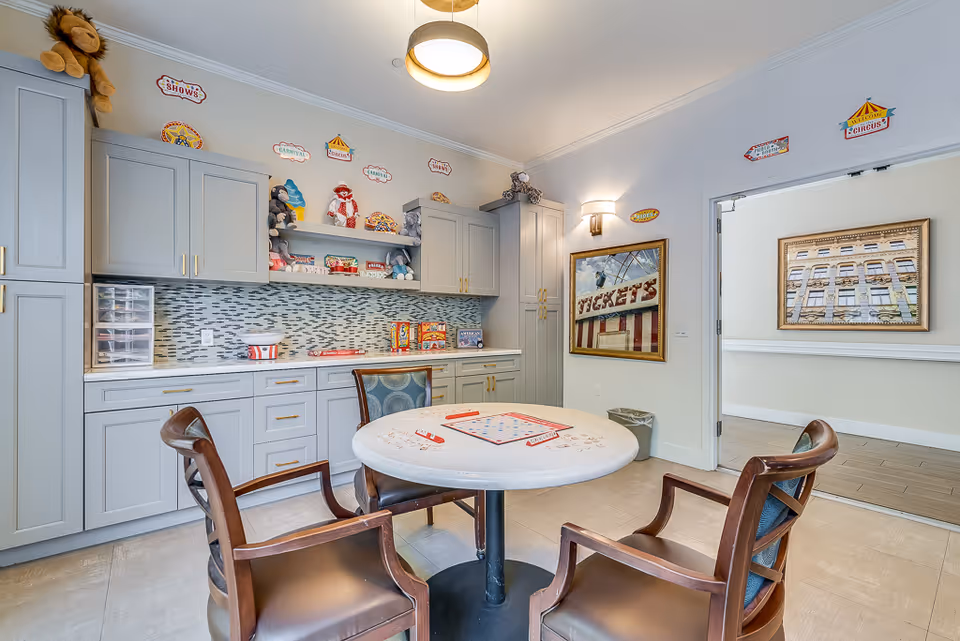 A cozy game room with a round table and four wooden chairs. The table has a Scrabble board game set up on it. The room features light gray cabinetry with gold handles, a mosaic tile backsplash, and shelves displaying stuffed animals and circus-themed decorations. There is a framed picture on the wall and a doorway leading to another room with a framed photo of a building.