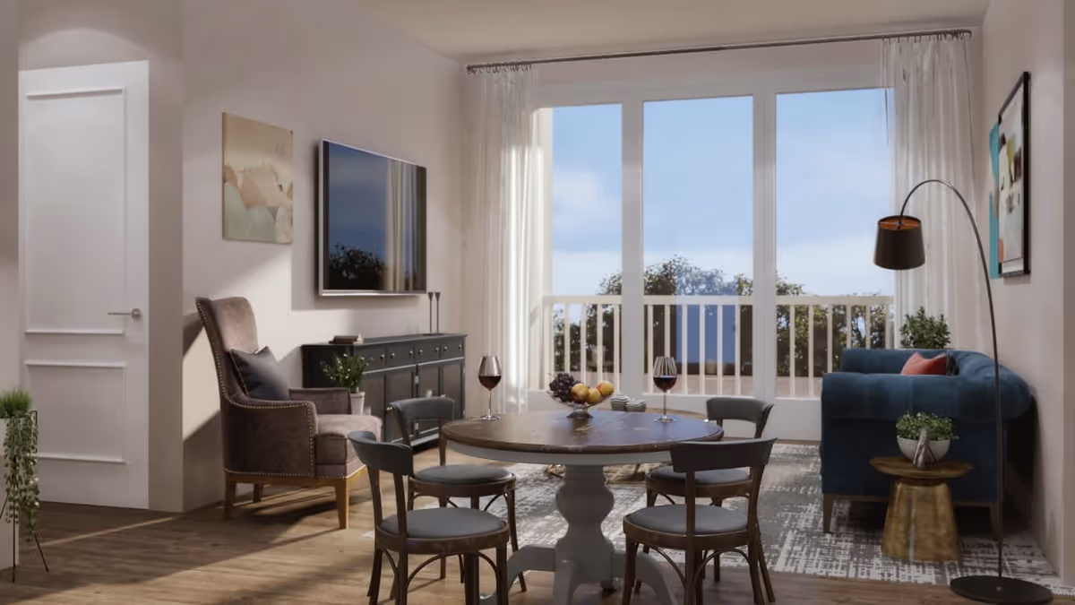 A bright living room with a round wooden table surrounded by four chairs, two glasses of red wine, and a bowl of fruit on the table. There is a blue sofa with a red pillow, a floor lamp, a small side table with a plant, and a large window with sheer curtains letting in natural light. A flat-screen TV is mounted on the wall above a black console cabinet, and a cushioned armchair is placed near the door.