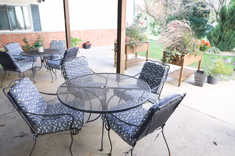 Outdoor patio area with metal tables and chairs featuring blue and white patterned cushions. There are potted plants and flower boxes with greenery and flowers around the patio, and a grassy area with trees in the background.