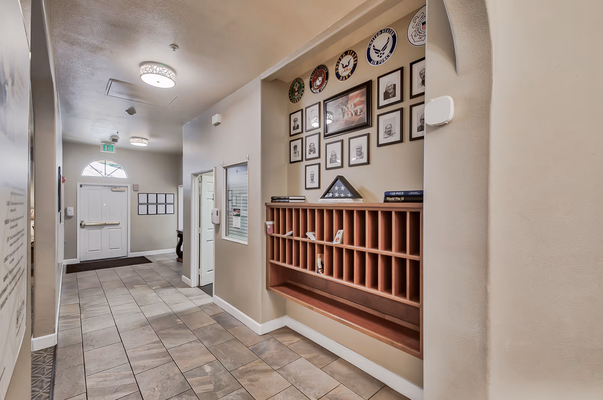 Interior hallway of Sunlit Gardens Assisted Living & Memory Care with tiled floor, beige walls, and ceiling lights. On the right wall, there is a wooden cubby mail organizer with framed photos and military insignias above it. A white door with a window and an exit door with an arched window above it are visible at the end of the hallway.
