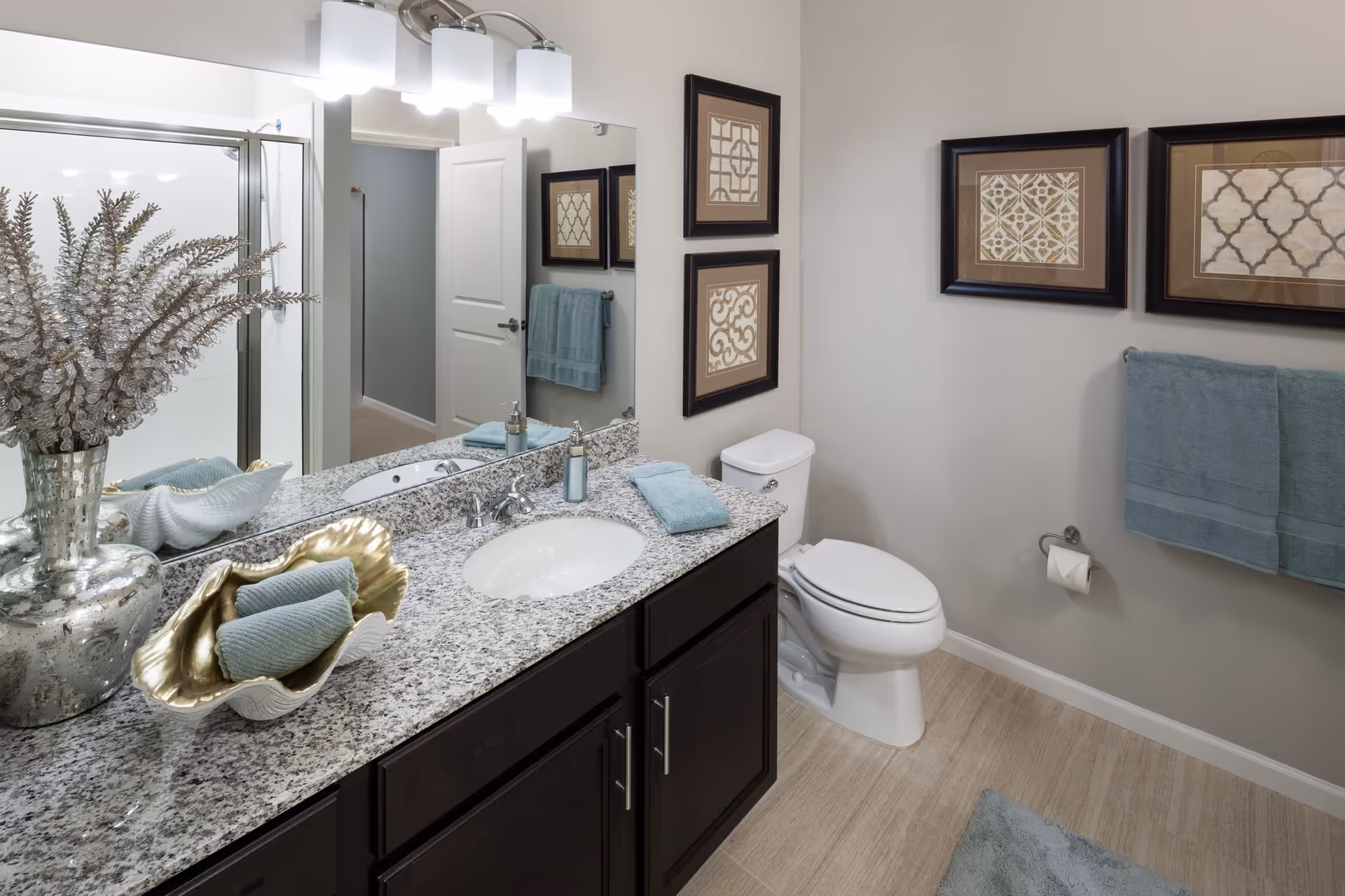 A modern bathroom with a granite countertop vanity featuring a sink, a silver vase with decorative branches, and neatly folded blue towels. The bathroom has a toilet, a glass-enclosed shower, three framed decorative artworks on the wall, and blue towels hanging on a towel rack. The floor has light-colored tiles and the walls are painted light gray.