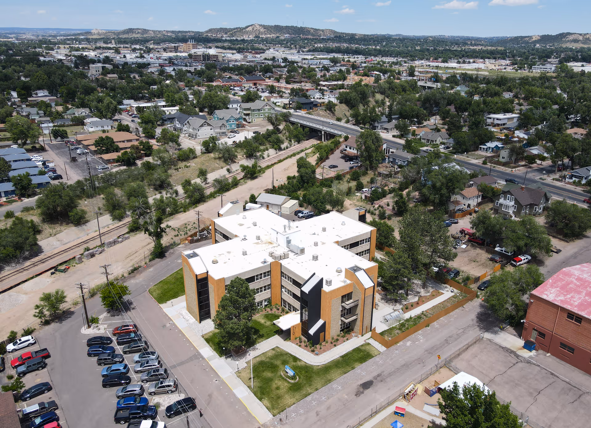 Aerial view of Springs Village Care Center, a multi-story building with a white roof and brown exterior, surrounded by parking lots, trees, and nearby residential and commercial areas under a clear blue sky.