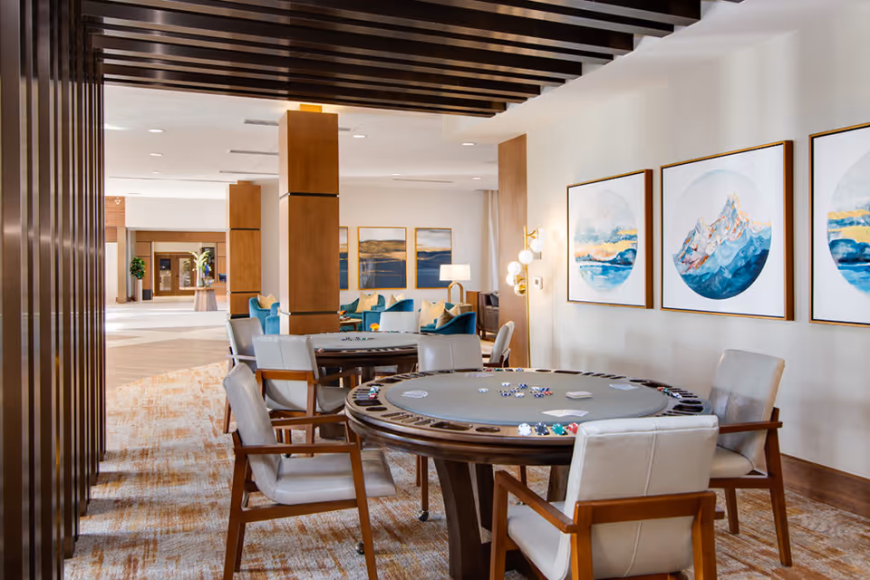 A bright and spacious common area in a senior living facility featuring a round poker table with chairs around it, poker chips and cards on the table, modern wall art depicting mountain and ocean scenes, and a seating area with blue and beige cushions in the background.