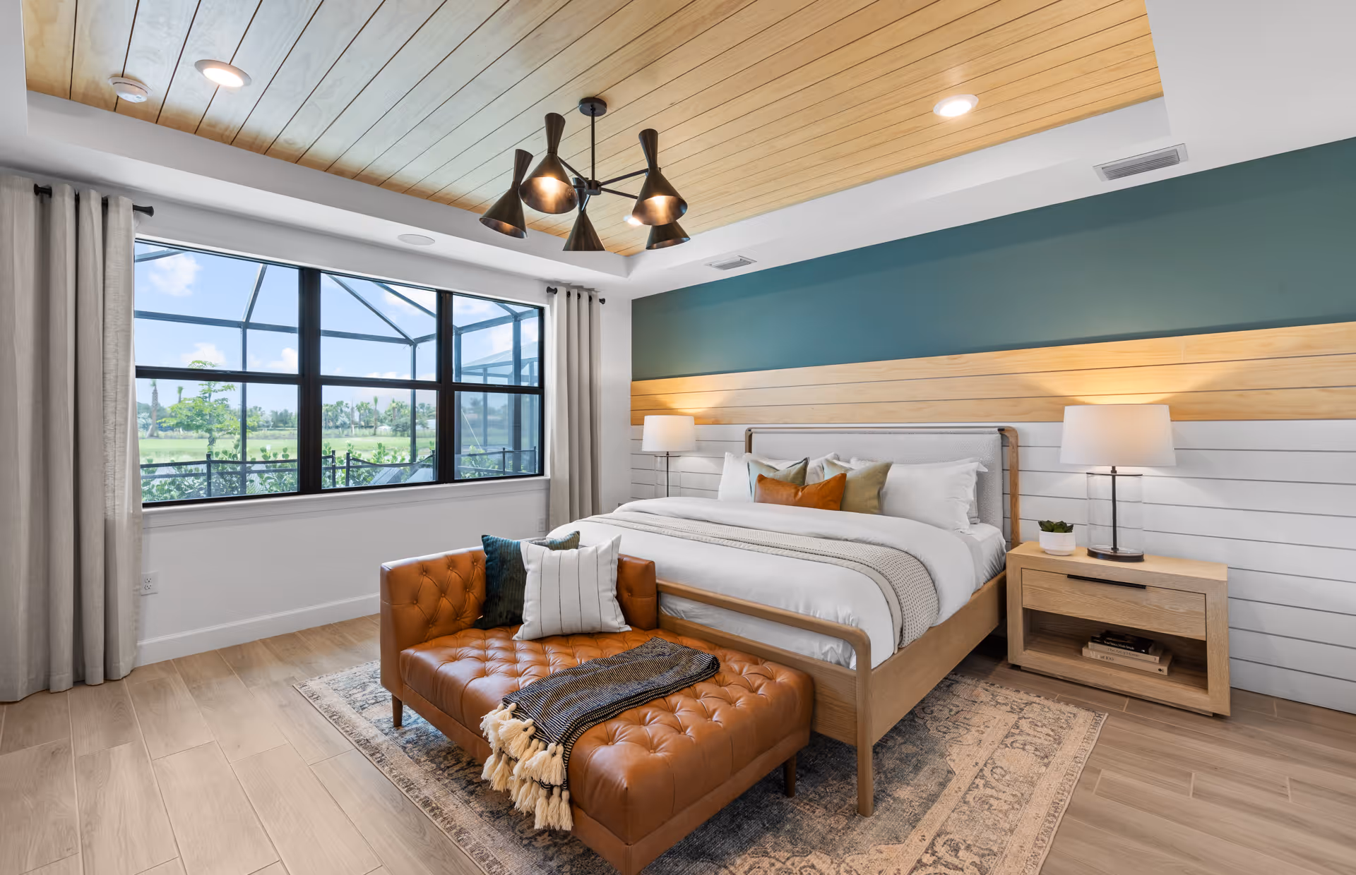 A modern bedroom with a large bed featuring white and beige bedding and multiple pillows. There is a brown tufted leather bench at the foot of the bed with a throw blanket and pillows. The room has a wooden ceiling with a contemporary black chandelier, light wood flooring, and a large window with beige curtains showing an outdoor view. Two bedside tables with lamps flank the bed, and the walls have a combination of teal, wood paneling, and white horizontal panels.