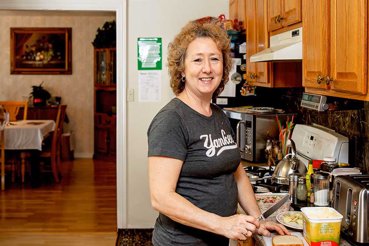 A smiling woman with curly hair wearing a Yankees t-shirt is preparing food in a kitchen. She is slicing bread on a countertop with various kitchen appliances and items around her. In the background, there is a dining area with a table and chairs.