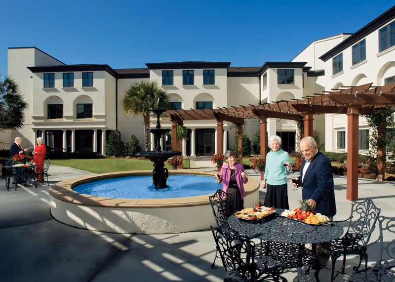 An outdoor courtyard area at Westminster Village featuring a circular water fountain in the center, surrounded by a patio with black wrought iron tables and chairs. Three elderly people stand near one table with trays of food and drinks, while another elderly couple is seated at a table in the background. The building has cream-colored walls with multiple windows and a wooden pergola providing partial shade.