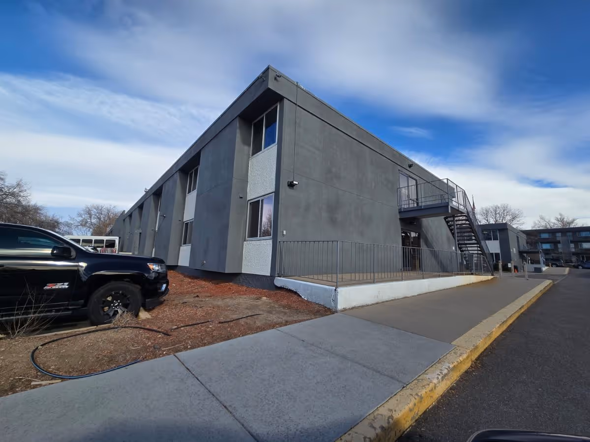 Exterior view of a gray two-story care facility with an outdoor staircase, sidewalk, and parked vehicles under a blue sky.