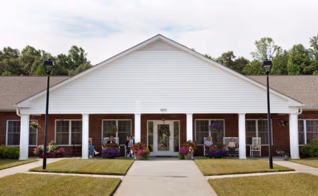 Single-story brick senior living facility front entrance with a covered porch, white columns, rocking chairs, and potted flowers.