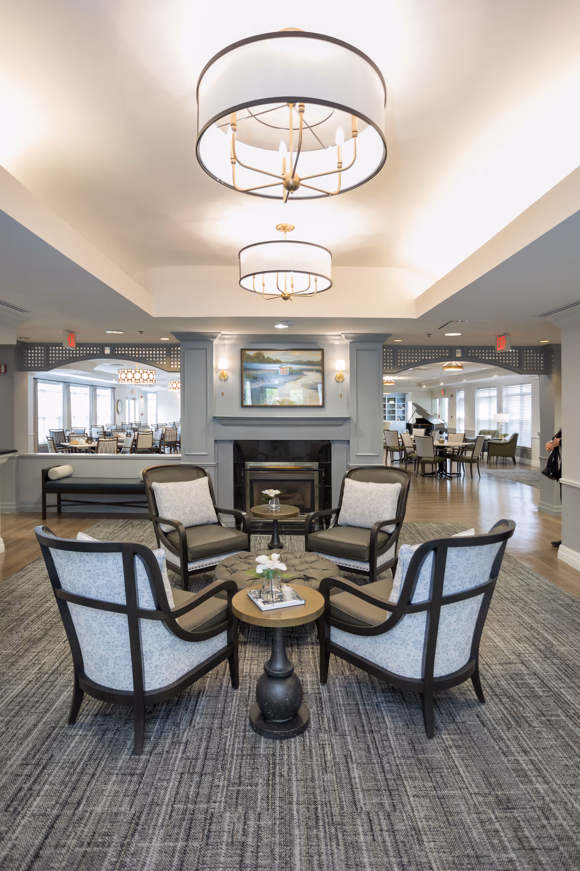 A bright communal seating area with four armchairs around a small table facing a fireplace, chandeliers overhead, and dining spaces visible in the background.
