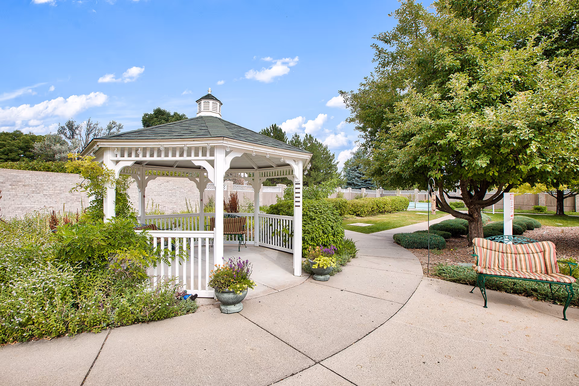 White gazebo in a landscaped courtyard with trees, potted plants, a striped bench and paved walkways.