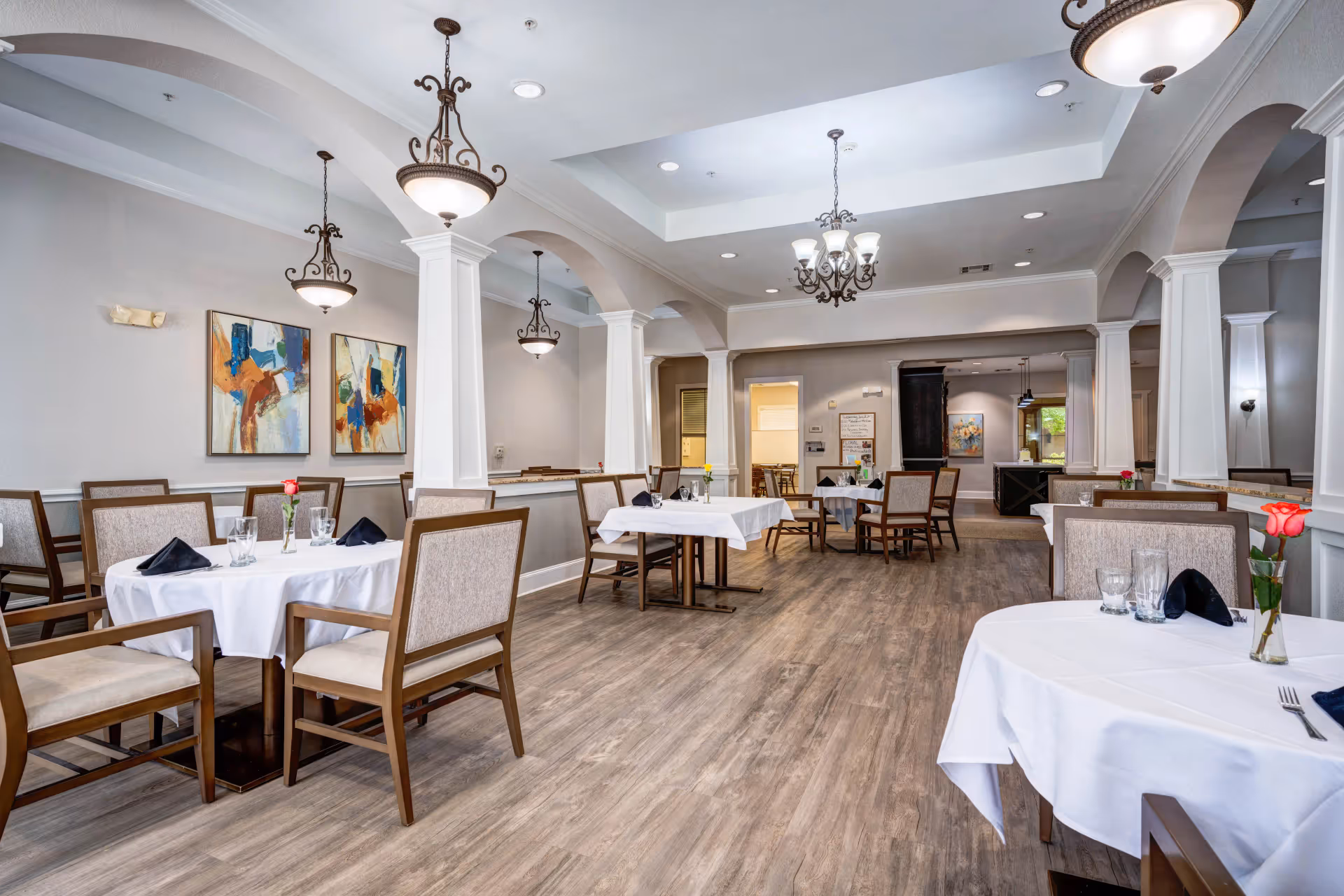 A spacious and elegant dining room with round tables covered in white tablecloths, each set with glasses, napkins, and a single rose in a vase. The room features wooden chairs with cushioned seats, decorative chandeliers hanging from the ceiling, and abstract artwork on the walls. The flooring is wood-style, and the room has white columns and arches adding to the sophisticated ambiance.