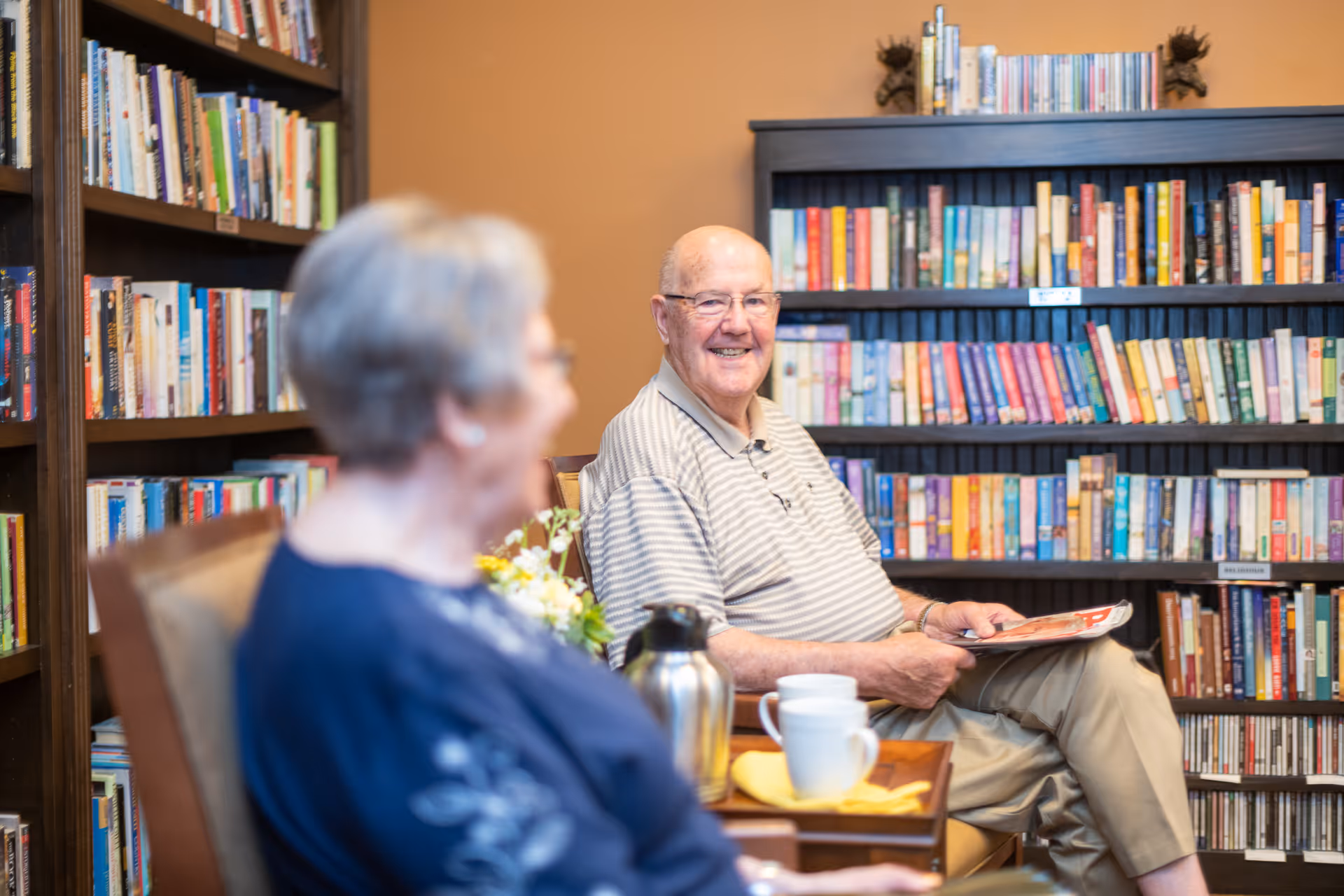 An elderly man and woman sitting in a cozy library or reading room with bookshelves filled with books behind them. The man is smiling and holding a magazine, while the woman is slightly out of focus in the foreground. There is a table with a coffee pot, cups, and a yellow napkin between them.