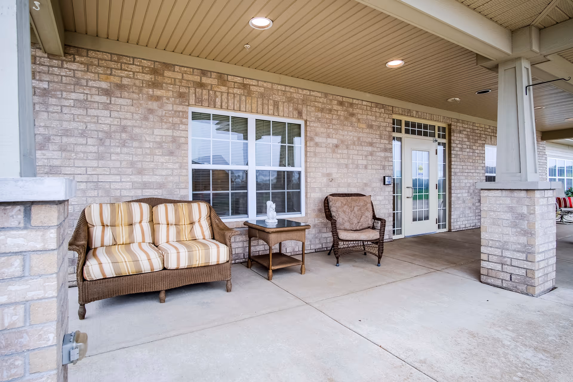 Covered outdoor patio area with a wicker loveseat with striped cushions, a wicker armchair with a patterned cushion, and a small wicker table with a decorative statue on it. The patio has a brick wall with windows and a glass door leading inside.