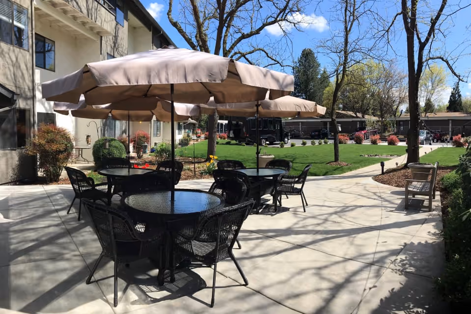 Outdoor courtyard with patio tables, umbrellas and chairs on a paved area beside a lawn and building.