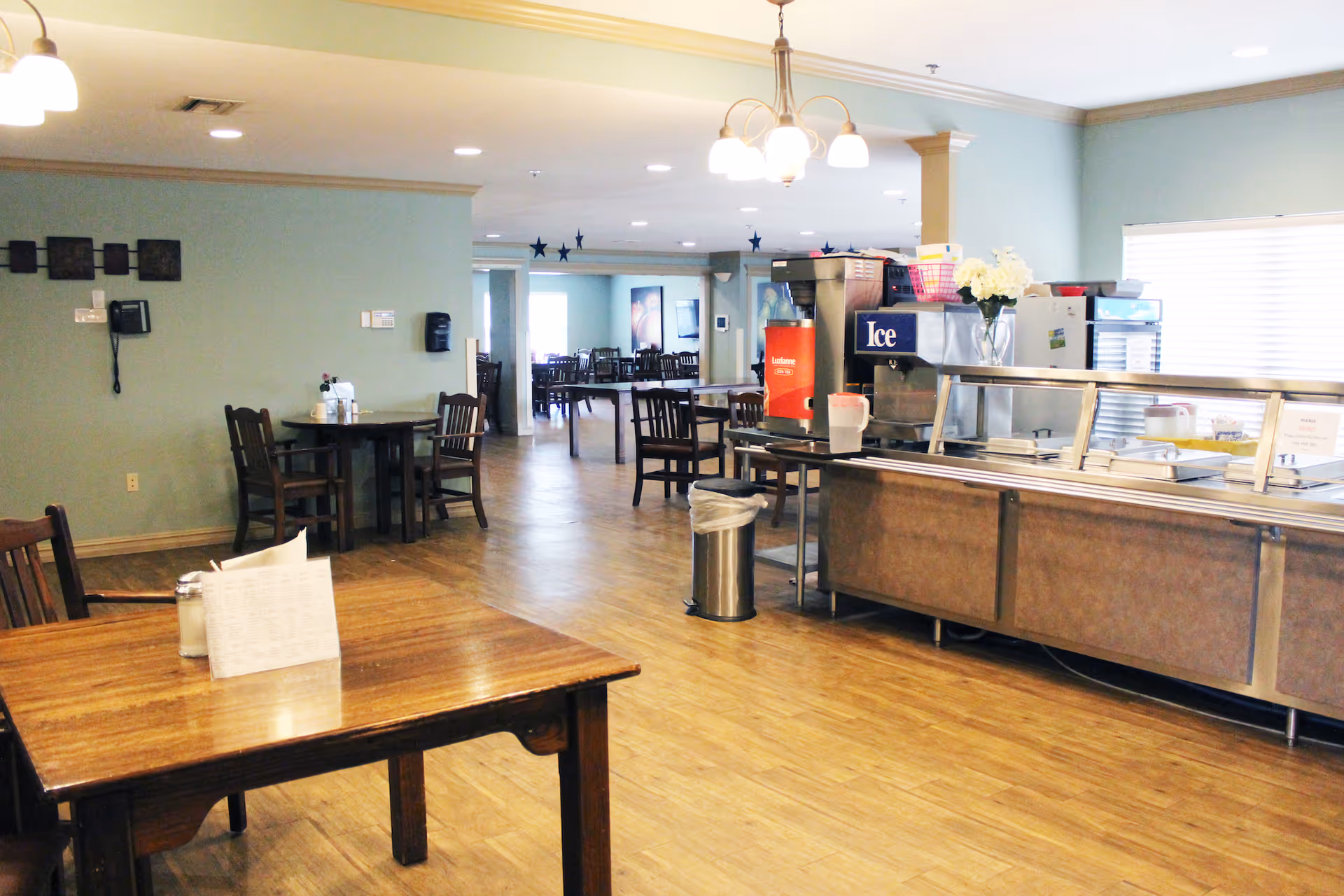 Dining room/cafeteria with a serving counter, ice machine, tables and chairs in a senior living facility.